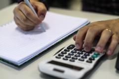 Imagem mostra uma pessoa fazendo cálculos em uma calculadora e anotando em um caderno.