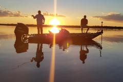 Barco a motor com dois pescadores pescando no por do sol, que se reflete na água
