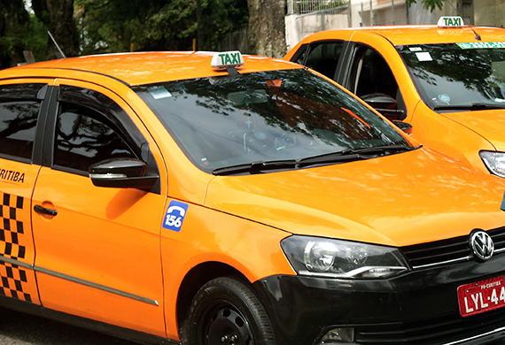 Visão parcial de um veículo de táxi de Curitiba na cor laranja pouco à frente de outro táxi