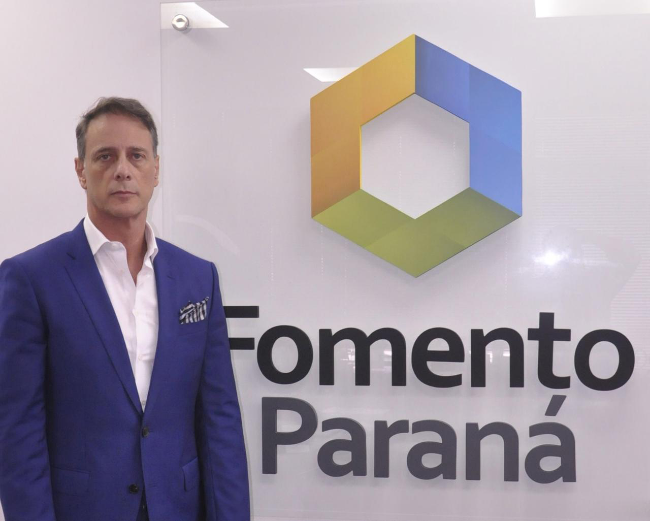 Claudio Stabile é um homem branco, veste terno azul e camisa branda, está à esquerda da imagem. Na parede aparece a dlogomarca da Fomento Paraná, um hexágono colorido com dois lados em azul, dois em amarelo e dois em verde. Abaixo está escrito Fomento Paraná.