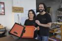 Patrícia Motta e Daniel Cortezia são jovens, brancos, de cabelos escuros, ele é mais alto e usa barba e cabelos compridos. Eles vestem camisetas pretas e mostram produtos da cartonagem que montaram com recursos da Fomento Paraná