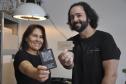 Patrícia Motta e Daniel Cortezia são jovens, brancos, de cabelos escuros, ele é mais alto e usa barba e cabelos compridos. Eles vestem camisetas pretas e mostram produtos da cartonagem que montaram com recursos da Fomento Paraná