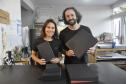 Patrícia Motta e Daniel Cortezia são jovens, brancos, de cabelos escuros, ele é mais alto e usa barba e cabelos compridos. Eles vestem camisetas pretas e mostram produtos da cartonagem que montaram com recursos da Fomento Paraná