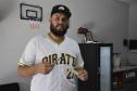 O empreendedor Brendon Oliveira é barbeiro ele usa camiseta branca com a palavra PIRATES e o número 21 estampado. Tem uma pente e tesoura nas mãos e está dentro da barbearia.