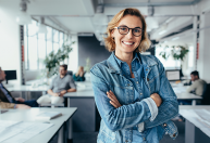 Mulher loira com jaqueta jeans azul de braços cruzados sorrindo em escritório com pessoas ao fundo, ambiente claro.