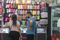 Mulher parda, de cabelos lisos até o meio das costas com camiseta rosa e calça preta ao lado de um homem pardo de cabelo curto e barba castanho escuro, de camiseta azul e calça preta. Os dois estão em frente um balcão olhando capinhas de celulares.