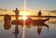 Pequeno barco a motor com dois pescadores pescando no por do sol, que se reflete na água, sem nenhum vento