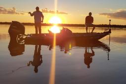 Pequeno barco a motor com dois pescadores pescando no por do sol, que se reflete na água, sem nenhum vento