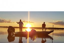 Dois homens pescando em um barco a motor parado em águas calmas. O sol está se pondo e a luz reflete na água passando pelo barco