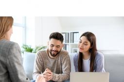 Um casal é atendido em casa por uma agente de crédito. Eles estão sentados em um sofá à frente de um computador. A agente está de costas