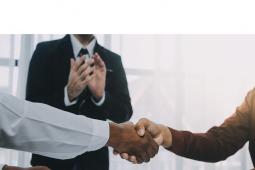 Duas pessoas apertando a mão simulando o fechamento de um negócio. Algém observa ao fundo, aplaudindo