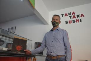 Ailton aparece em primeiro plano segurando embalagem preta com logo de seu restaurante. Atrás dele está escrito "Taka Taka Sushi"