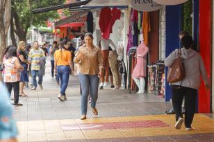 Calçada com pessoas passando na frente de diversas lojas, com manequins e roupas penduras na entradada dos comércios