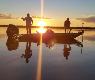 Pequeno barco a motor com dois pescadores pescando no por do sol, que se reflete na água, sem nenhum vento