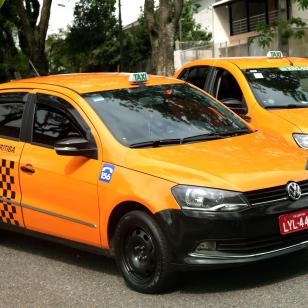 Visão parcial de um veículo de táxi de Curitiba na cor laranja pouco à frente de outro táxi