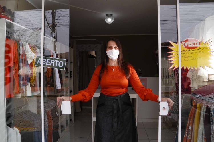 A empresária Josiane Duarte, proprietária de uma loja de roupas para o público evangélico, em Piraquara. Na foto ela está na porta da loja, em vidro, com placa de "aberto", e dá para ver as roupas atrás dela.