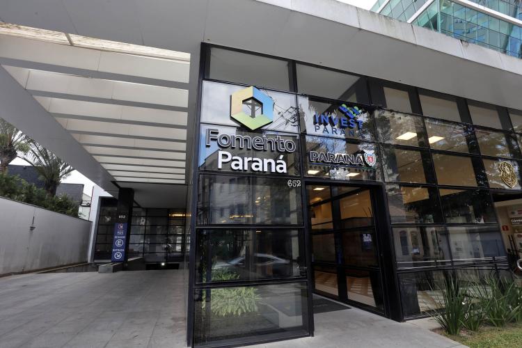 Imagem da fachada da sede da Fomento Paraná em Curitiba. É uma parede envidraçada em módulos tipo fumê com logotipos da Fomento, Invest e Governo