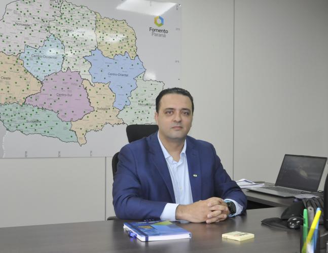 Gustavo Emanuel Cejas é um homem branco, usa terno azul escuro e camisa azul clara. Está sentado em uma mesa de escritório. Há um mapa do Paraná na parede atrás dele e um lap top na mesa ao lado.