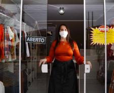 A empresária Josiane Duarte, proprietária de uma loja de roupas para o público evangélico, em Piraquara. Na foto ela está na porta da loja, em vidro, com placa de "aberto", e dá para ver as roupas atrás dela.