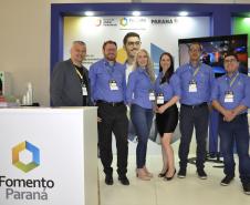 Equipe Fomento Paraná, no atendimento aos empresários e dirigentes de associações comerciais e empresariais. Grupo de seis pessoas, com camisas azuis, em pé, ao lado de um balcão, no estande da Fomento Paraná no evento.