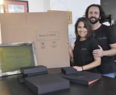 Patrícia Motta e Daniel Cortezia são jovens, brancos, de cabelos escuros, ele é mais alto e usa barba e cabelos compridos. Eles vestem camisetas pretas e mostram produtos da cartonagem que montaram com recursos da Fomento Paraná