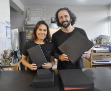 Patrícia Motta e Daniel Cortezia são jovens, brancos, de cabelos escuros, ele é mais alto e usa barba e cabelos compridos. Eles vestem camisetas pretas e mostram produtos da cartonagem que montaram com recursos da Fomento Paraná