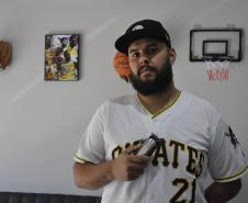 O empreendedor Brendon Oliveira é barbeiro ele usa camiseta branca com a palavra PIRATES e o número 21 estampado. Tem uma máquina de cortar cabelos na mão direita e na parede, atrás dele,tem uma cesta de basquete, um quadro com jogadores negros e uma luva de basebal