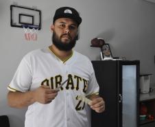 O empreendedor Brendon Oliveira é barbeiro ele usa camiseta branca com a palavra PIRATES e o número 21 estampado. Tem uma pente e tesoura nas mãos e está dentro da barbearia.