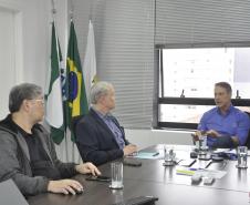 Claudio Stabile, de camisa azul claro, à direita, conversa com Jorge Callado e Julio Suzuki, todos sentados. Há uma janela aberta atrás do Claudio, sobre a mesa à copos de água e alguns papéis.