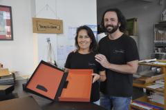 Patrícia Motta e Daniel Cortezia são jovens, brancos, de cabelos escuros, ele é mais alto e usa barba e cabelos compridos. Eles vestem camisetas pretas e mostram produtos da cartonagem que montaram com recursos da Fomento Paraná