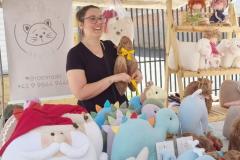 Uma mulher branca com cabelo castanho claro e óculos sorri enquanto segura uma pelúcia de capivara. Ela está atrás do balcão de uma barraca, que exibe várias pelúcias coloridas e diferentes em exibição.