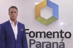 Claudio Stabile é um homem branco, veste terno azul e camisa branda, está à esquerda da imagem. Na parede aparece a dlogomarca da Fomento Paraná, um hexágono colorido com dois lados em azul, dois em amarelo e dois em verde. Abaixo está escrito Fomento Paraná.