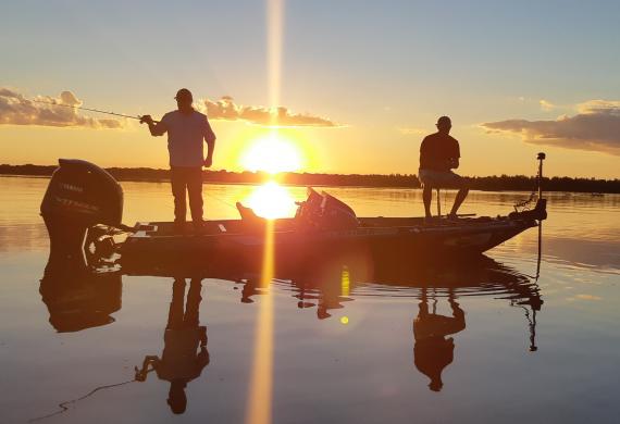 Pequeno barco a motor com dois pescadores pescando no por do sol, que se reflete na água, sem nenhum vento