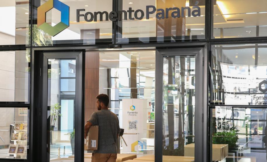 Na imagem, fachada da sede da Fomento Paraná em Curitiba. Há portas e janelas de vidro e o logotipo da instituição está fixado bem acima da entrada do prédio. Ao centro da fotografia, homem caminha em direção à recepção do edifício.