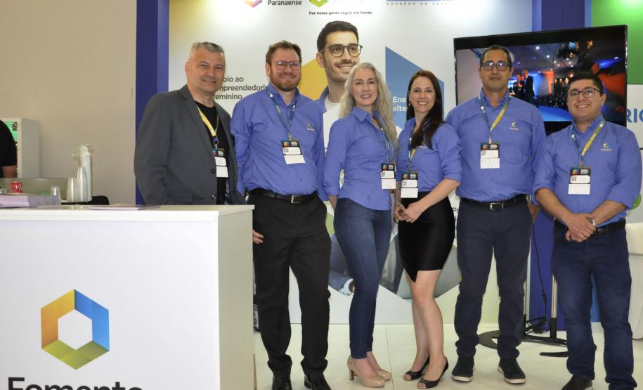 Equipe Fomento Paraná, no atendimento aos empresários e dirigentes de associações comerciais e empresariais. Grupo de seis pessoas, com camisas azuis, em pé, ao lado de um balcão, no estande da Fomento Paraná no evento.