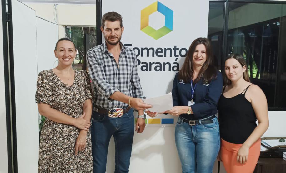 Imagem de quadro pessoas em pé com um papel na mão do empresário Evandro. Na parede ao fundo há um banner com a logomarca e o nome Fomento Paraná. Na imagem estão A secretária municipal de Indústria e Comércio de Francisco Beltrão, Liliana Andrade, com o empresário Evandro Bellin da Silva, a agente de crédito Luciana Dani e a estagiária Vitória Gallina.