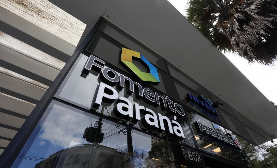 Parede envidraçada da fachada do edifício sede da Fomento Paraná, mostra logomarca da instituição em primeiro plano, formada por um hexágono nas cores azul, amarelo e verde.