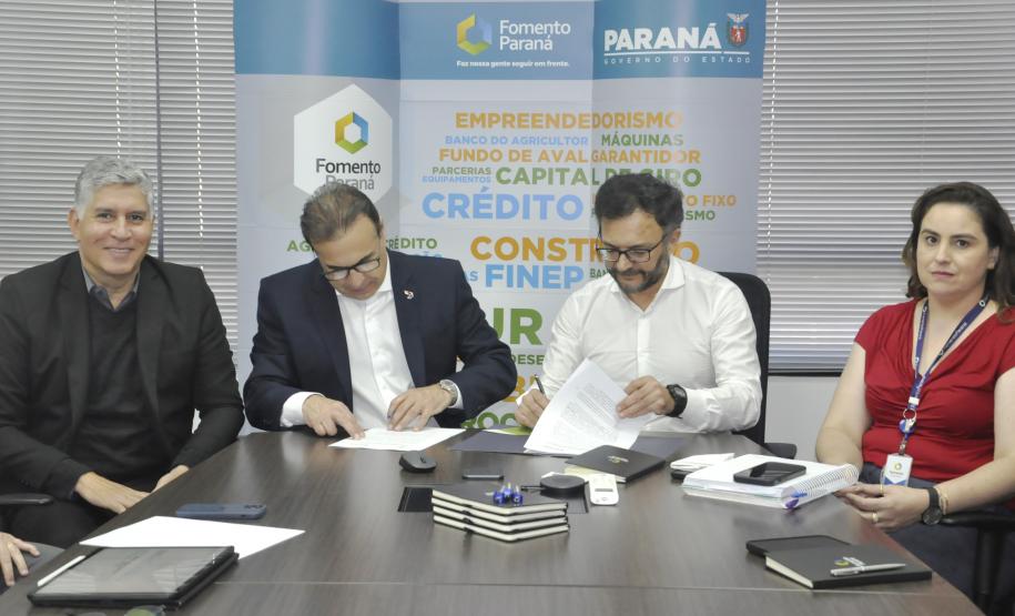 Reunião entre diretores da Fomento paraná e do BDMG em uma sala com uma grande mesa de reuniões. Presidentes da sduas instituições aqssinam documento.