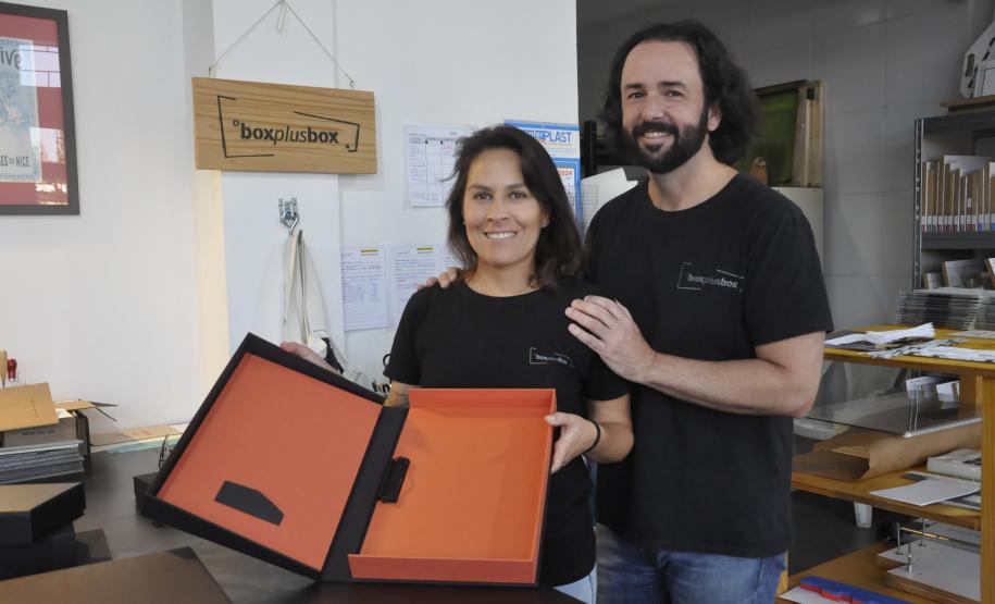 Patrícia Motta e Daniel Cortezia são jovens, brancos, de cabelos escuros, ele é mais alto e usa barba e cabelos compridos. Eles vestem camisetas pretas e mostram produtos da cartonagem que montaram com recursos da Fomento Paraná