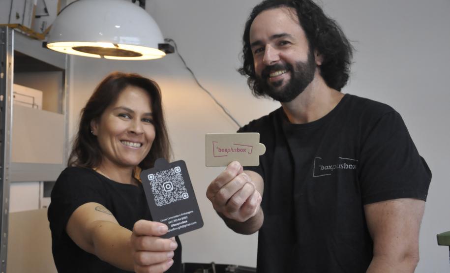 Patrícia Motta e Daniel Cortezia são jovens, brancos, de cabelos escuros, ele é mais alto e usa barba e cabelos compridos. Eles vestem camisetas pretas e mostram produtos da cartonagem que montaram com recursos da Fomento Paraná