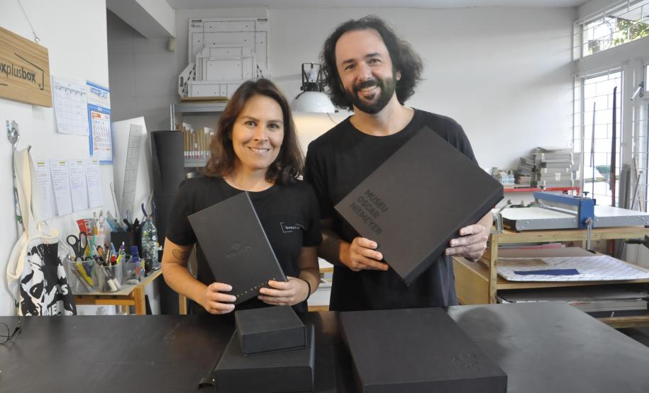 Patrícia Motta e Daniel Cortezia são jovens, brancos, de cabelos escuros, ele é mais alto e usa barba e cabelos compridos. Eles vestem camisetas pretas e mostram produtos da cartonagem que montaram com recursos da Fomento Paraná