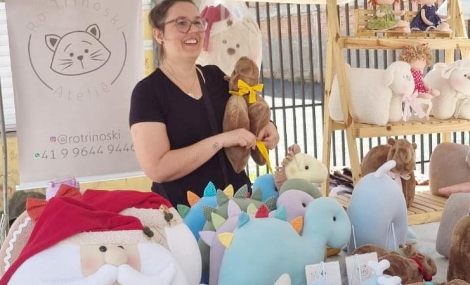 Uma mulher branca com cabelo castanho claro e óculos sorri enquanto segura uma pelúcia de capivara. Ela está atrás do balcão de uma barraca, que exibe várias pelúcias coloridas e diferentes em exibição.