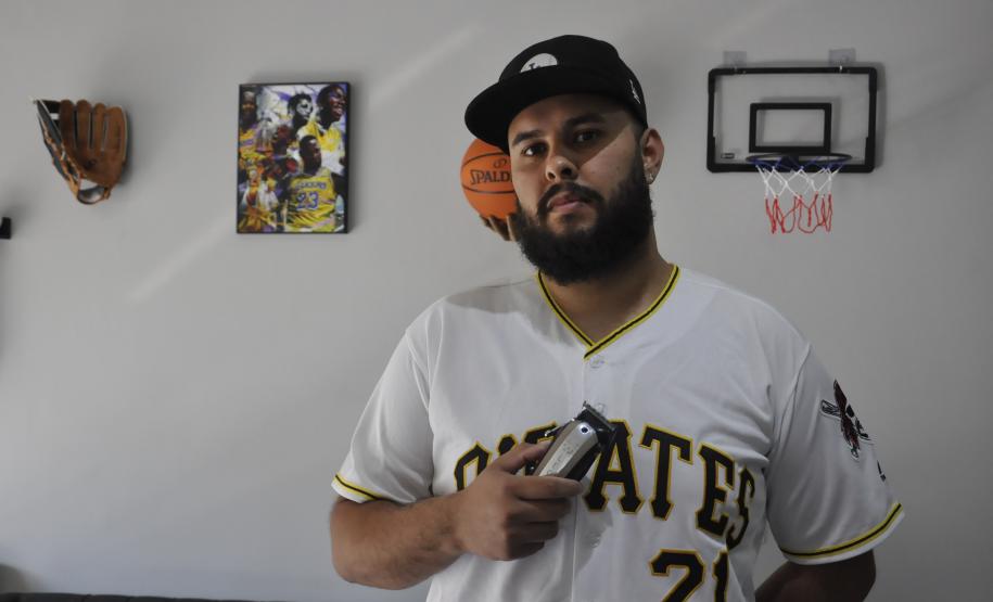 O empreendedor Brendon Oliveira é barbeiro ele usa camiseta branca com a palavra PIRATES e o número 21 estampado. Tem uma máquina de cortar cabelos na mão direita e na parede, atrás dele,tem uma cesta de basquete, um quadro com jogadores negros e uma luva de basebal