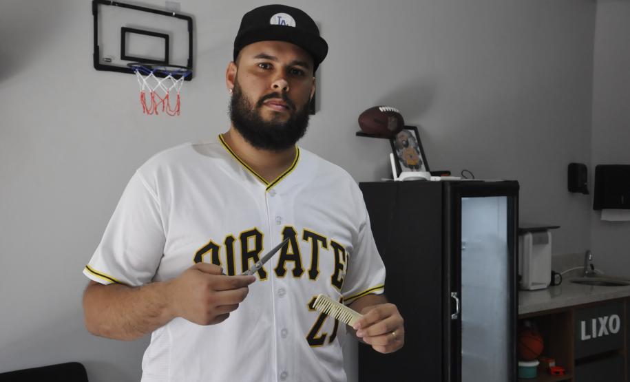 O empreendedor Brendon Oliveira é barbeiro ele usa camiseta branca com a palavra PIRATES e o número 21 estampado. Tem uma pente e tesoura nas mãos e está dentro da barbearia.