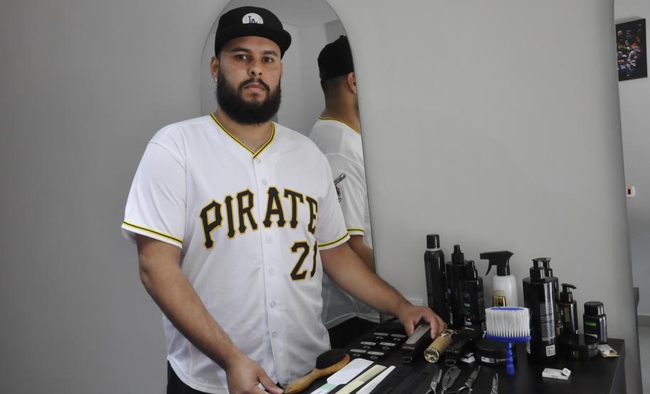 O empreendedor Brendon Oliveira é barbeiro ele usa camiseta branca com a palavra PIRATES e o número 21 estampado. Ele mostra as ferramentas de trabalho e produtos que usa na atividade dele.