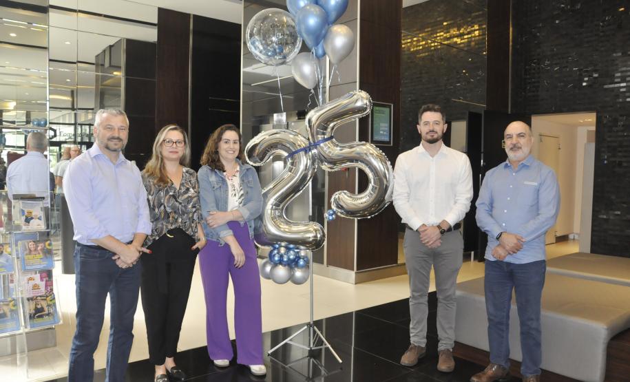 Diretores da Fomento Paraná posam para foto para marcar a comemoração de 25 anos. São duas mulheres, três homens, e no meio deles há um balão prateado no formato do número 25, preso a um tripé.