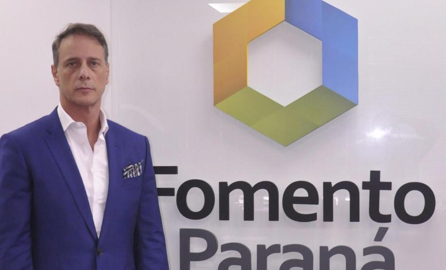 Claudio Stabile é um homem branco, veste terno azul e camisa branda, está à esquerda da imagem. Na parede aparece a dlogomarca da Fomento Paraná, um hexágono colorido com dois lados em azul, dois em amarelo e dois em verde. Abaixo está escrito Fomento Paraná.