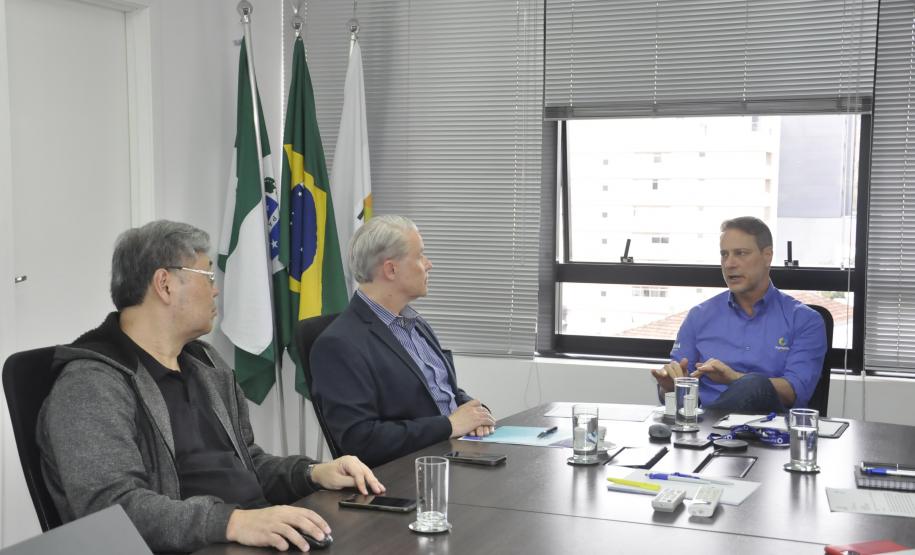 Claudio Stabile, de camisa azul claro, à direita, conversa com Jorge Callado e Julio Suzuki, todos sentados. Há uma janela aberta atrás do Claudio, sobre a mesa à copos de água e alguns papéis.