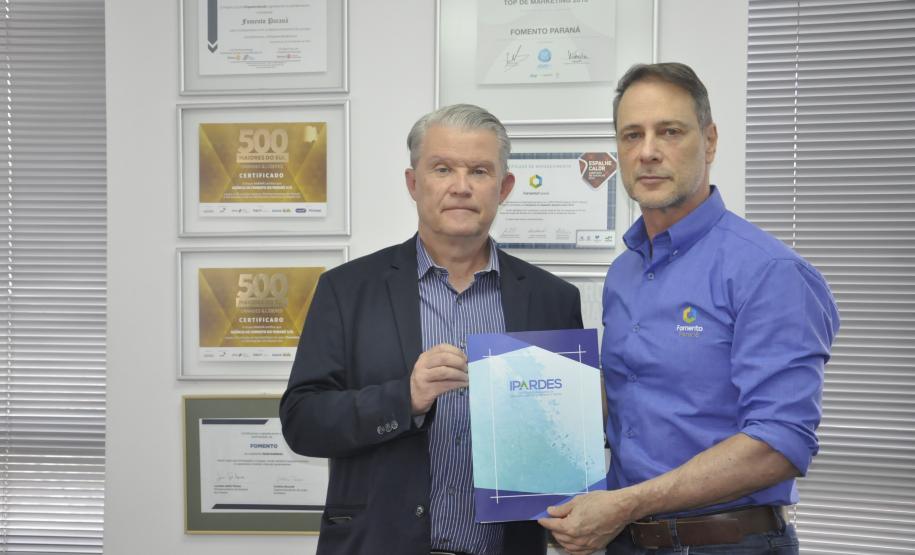 Dois homens brancos, Claudio Stabile, de camisa azul claro, à direita, e Jorge Callado, posam apresentando uma pasta de papel com uma iscrição na capa com o nome IPARDES, presidido por Callado.