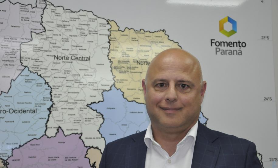 Claudio Pacheco é um homem branco, calvo, usa camisa branca e paletó azul marinho, tem ao fundo um mapara o Paraná com uma logomarca da Fomento Paraná